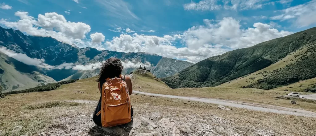 Autora sentada em colina descampada observa a igreja de Kazbegi, durante uma viagem para a Geórgia.